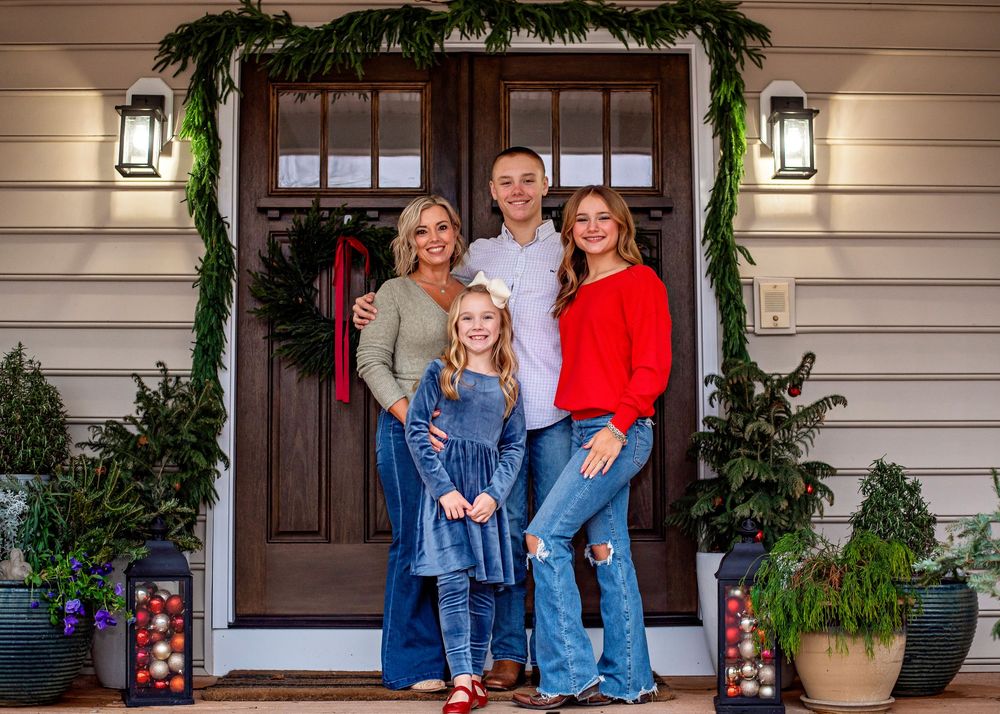 People wearing denim jackets and jeans sit together on a decorated front porch with holiday greenery and lanterns.