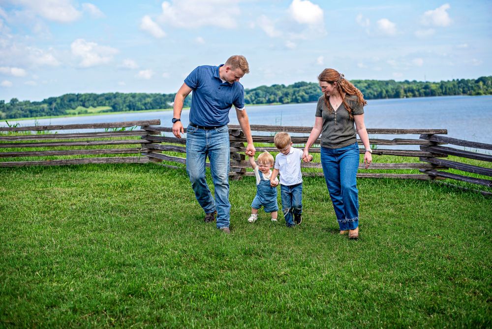Family of four walking on green grass near a wooden fence and lake on a sunny day.