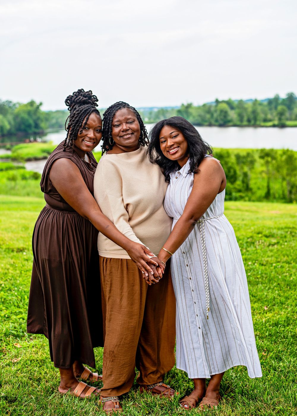Three women smiling and holding hands outdoors near a lake, wearing brown, beige, and white dresses on green grass.