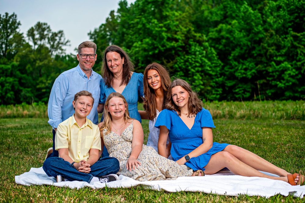 Happy family of six posing outdoors on a blanket in a green park, smiling for a portrait photo.