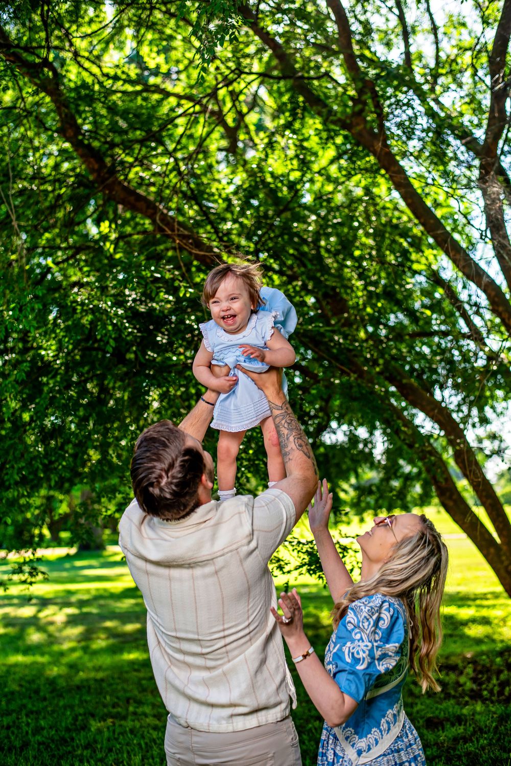 Parents joyfully lifting their laughing baby girl in a sunny green park, surrounded by lush trees.