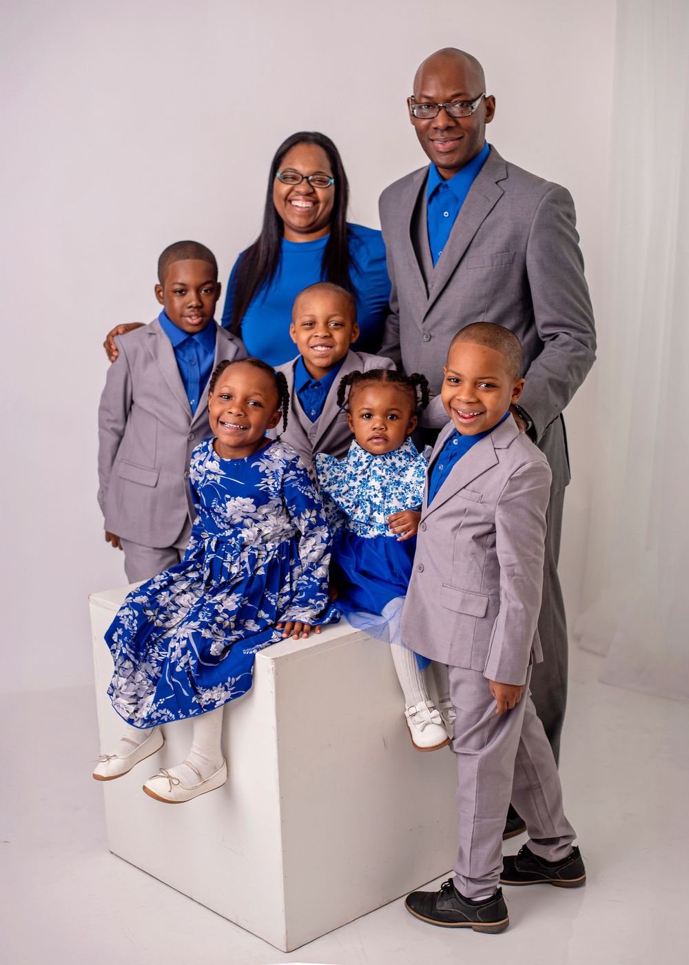 A family of six wearing coordinated blue and grey outfits poses together for a formal portrait against a white backdrop.