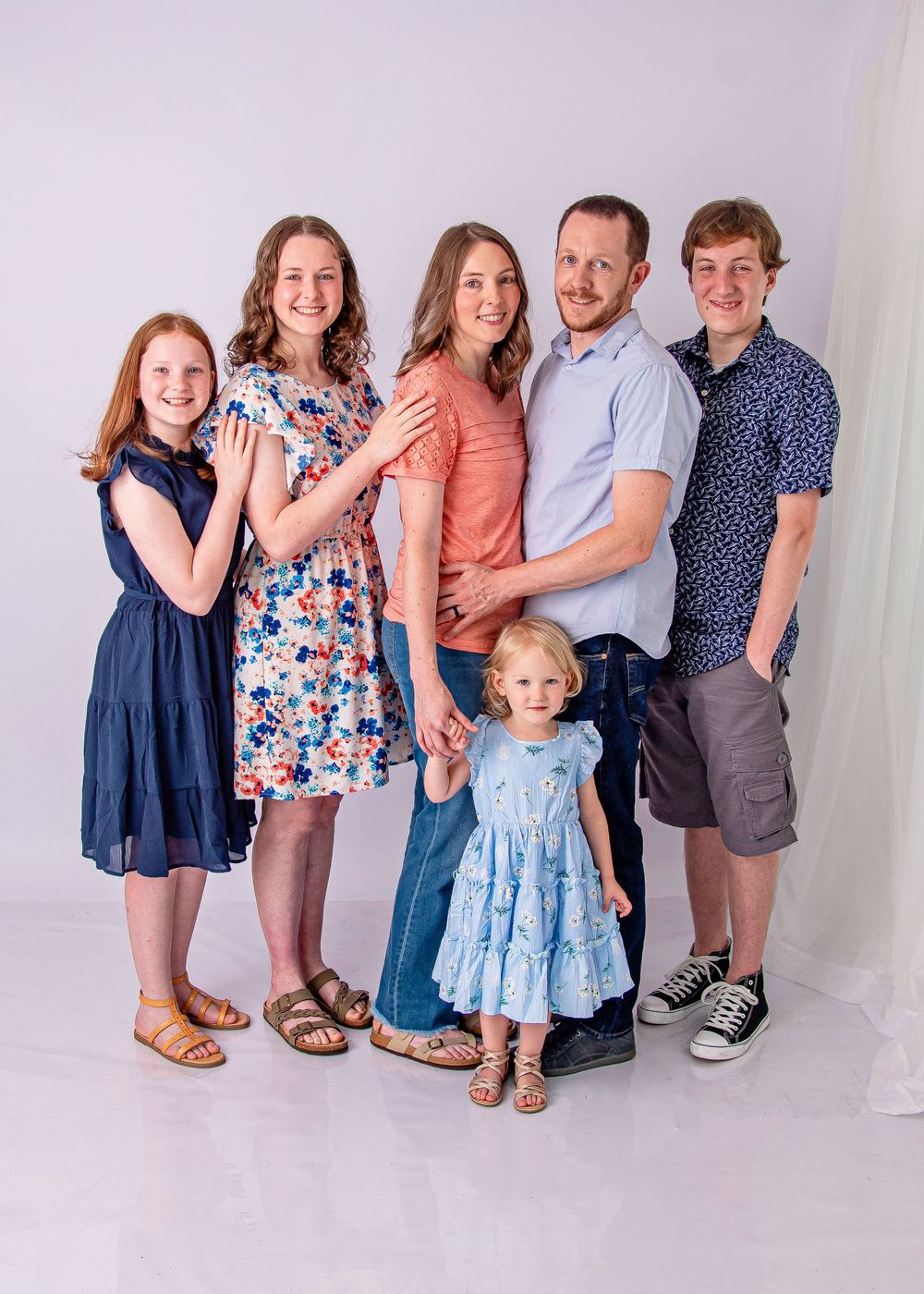 A joyful family of six poses together in a bright studio, including parents, three girls, and a teenage boy.