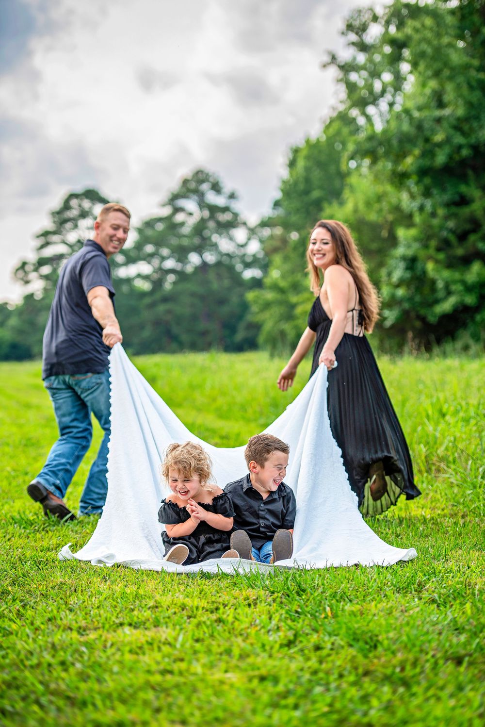 Family of four playing outdoors, parents holding white blanket over two young children sitting on lush green grass.