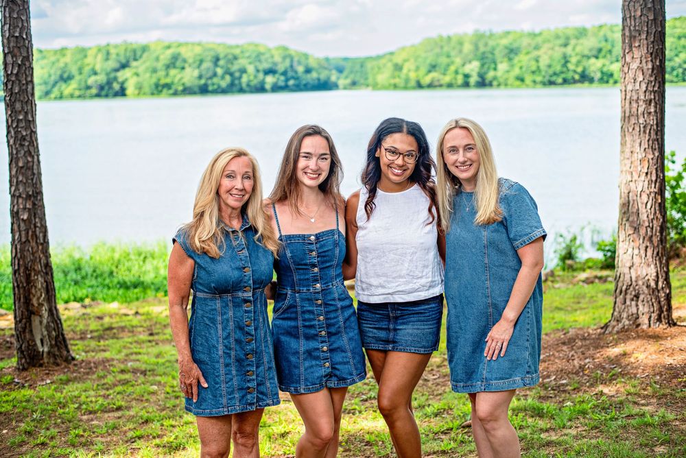 Four women smiling outdoors near a lake, wearing denim outfits, surrounded by trees and lush greenery.