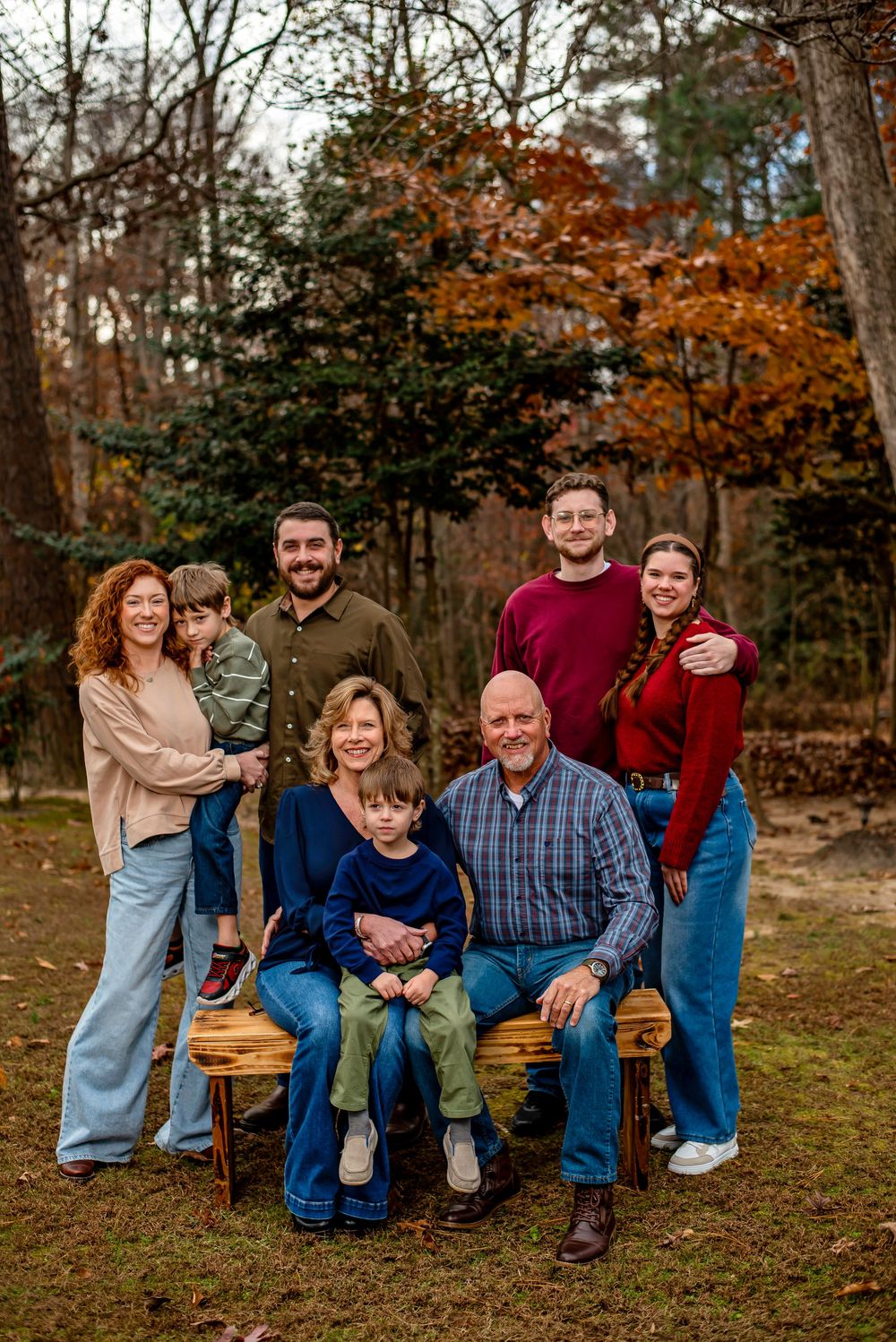 A family group poses together outdoors on a fall day with autumn foliage and trees in the background.