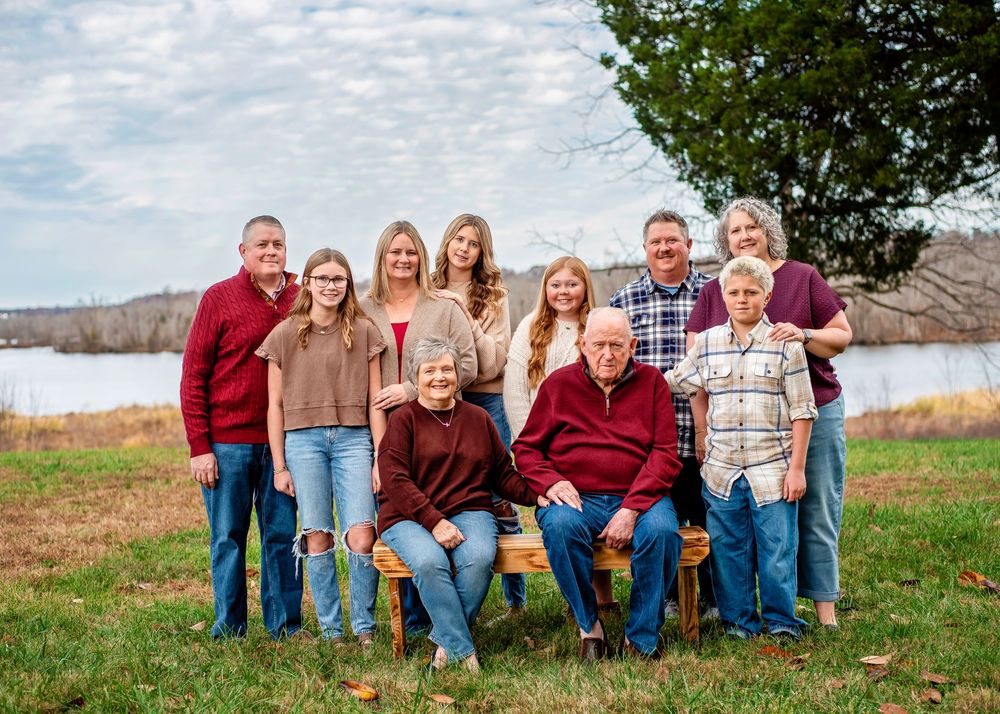 A family group sits together outdoors on wooden chairs against a scenic rural backdrop in coordinating outfits.