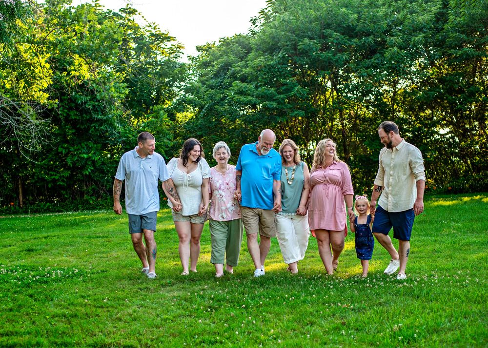 Multi-generational family walking together on green grass lawn surrounded by lush trees on a sunny day.