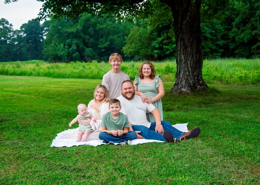A family poses together on a blanket under a large tree in a peaceful green summer meadow.
