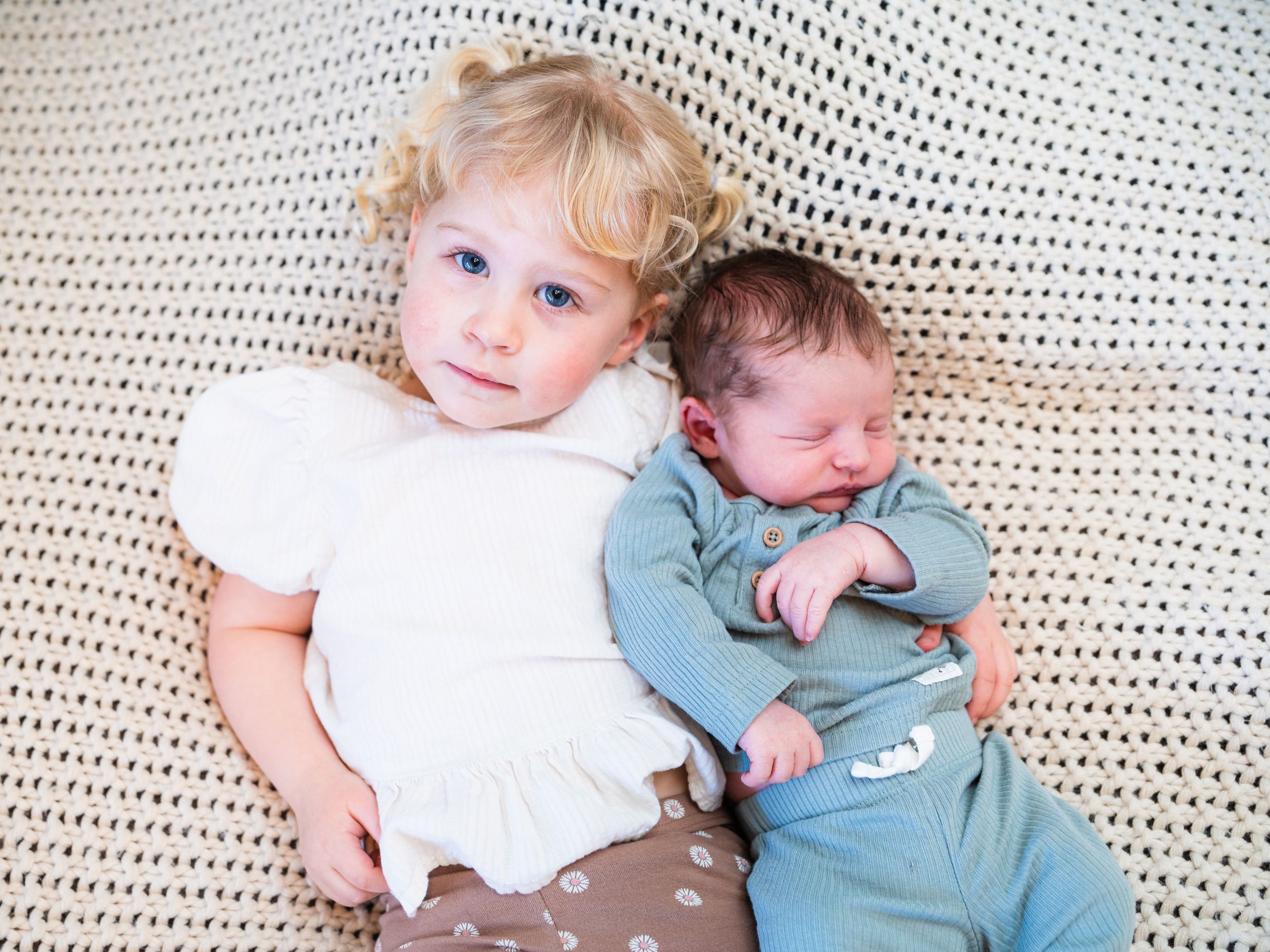 Two young children lying on a white knitted blanket, one wearing white and brown, the other in light blue during at home newborn sesion.