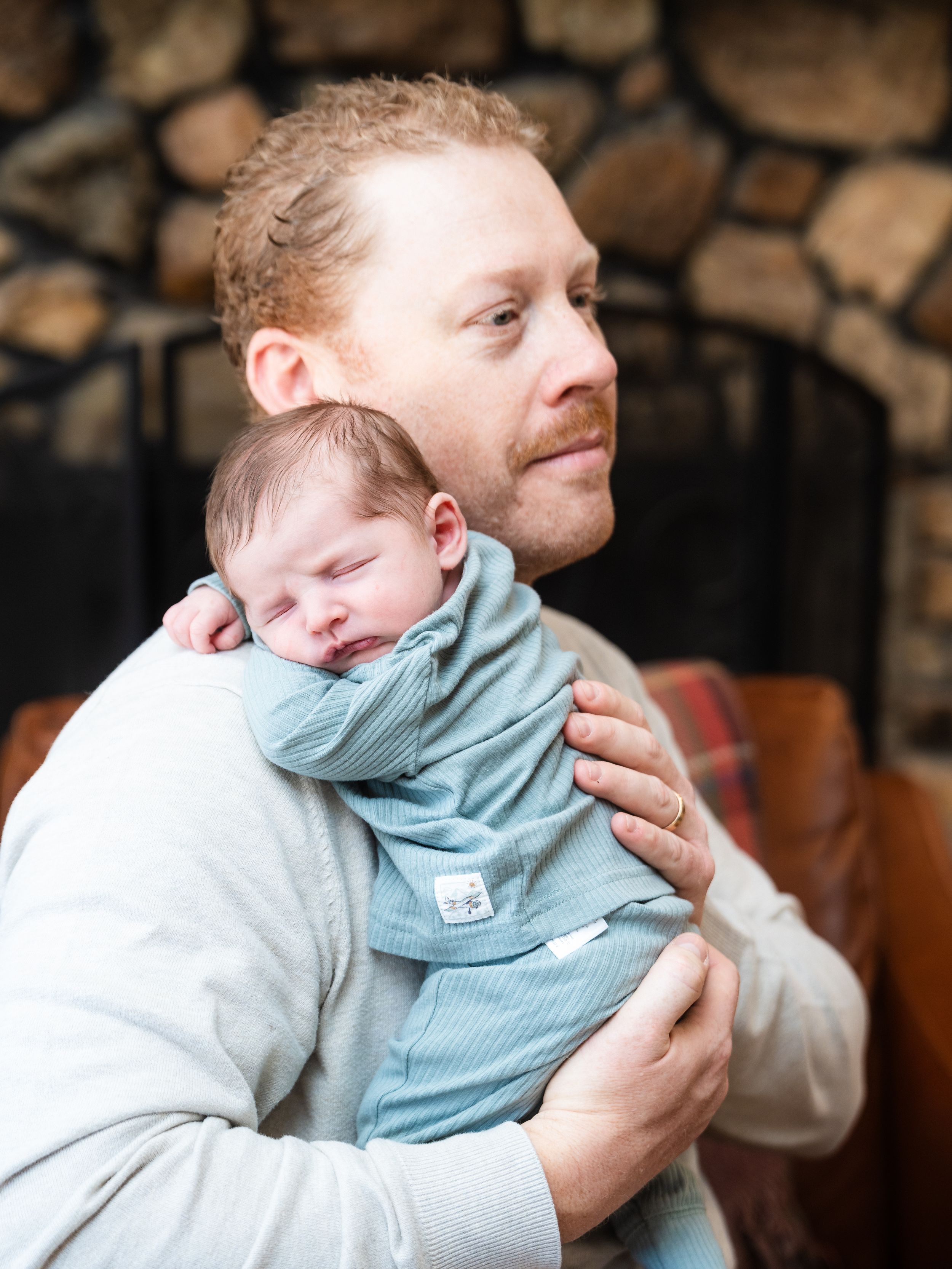 A sleeping baby in a light blue outfit rests peacefully while being held by his dad against a stone wall background.