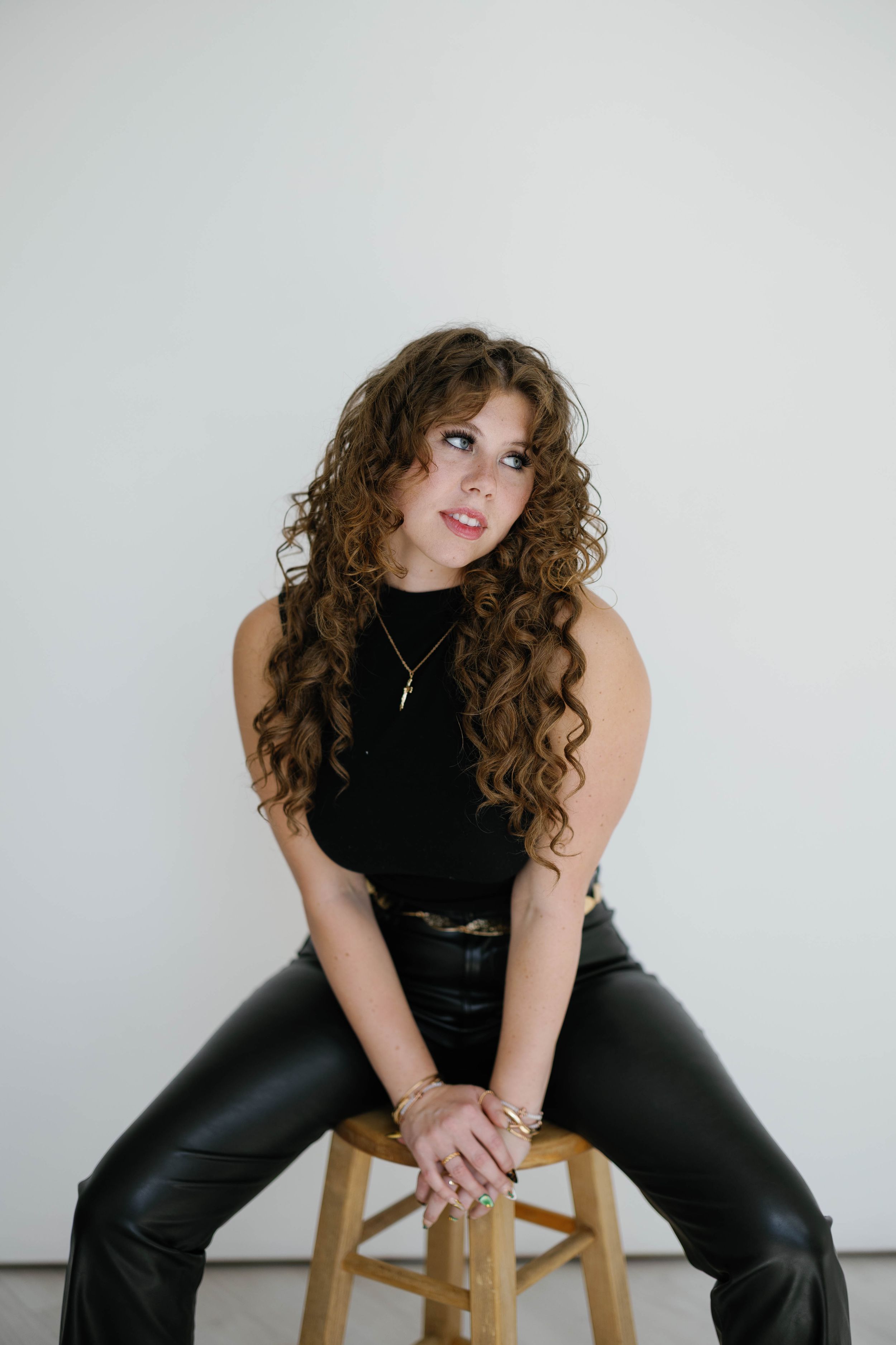 Young woman with long curly hair poses on wooden stool in black outfit against white wall.