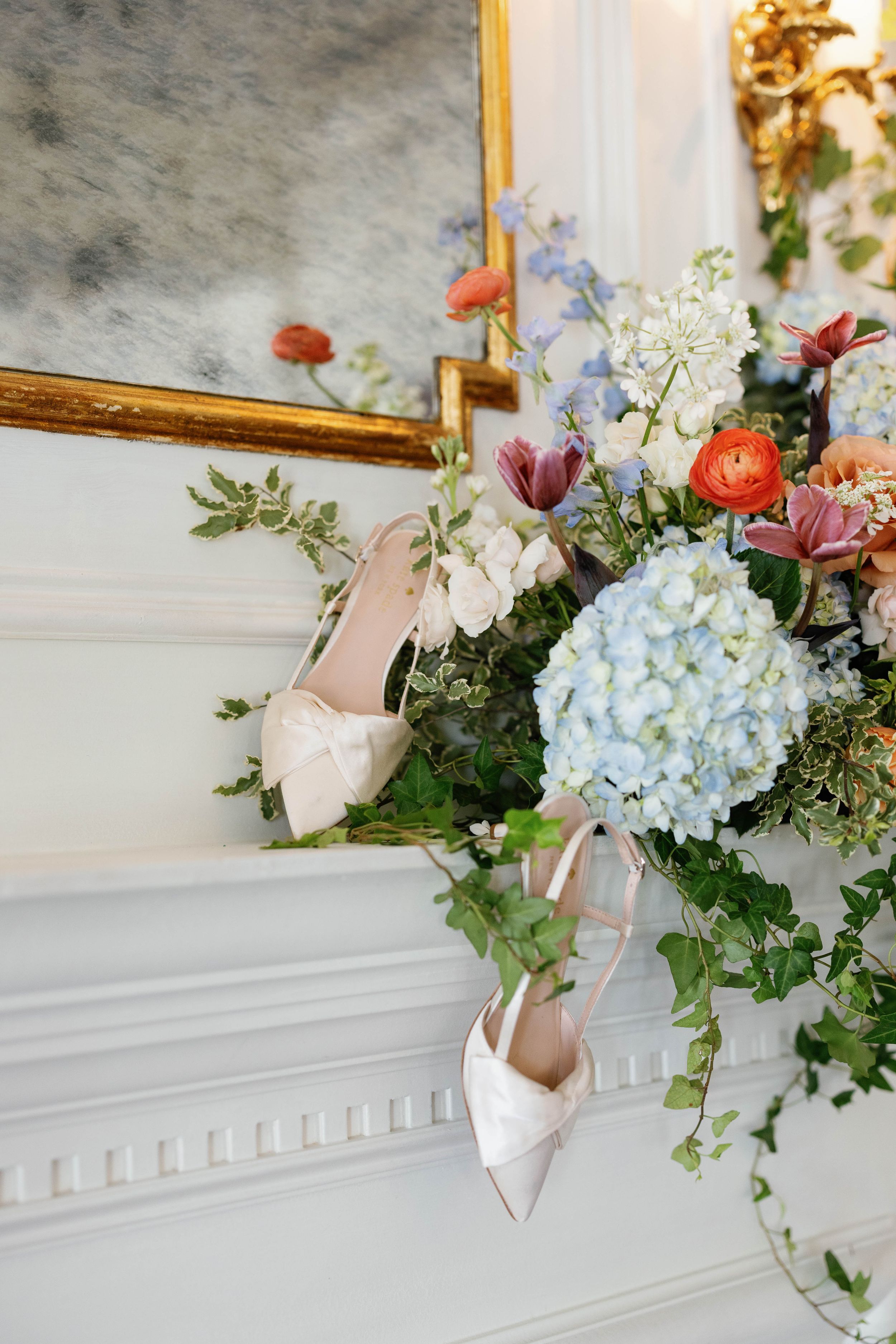 Elegant bridal heels resting on a white fireplace mantel adorned with colorful spring flowers including blue hydrangeas.