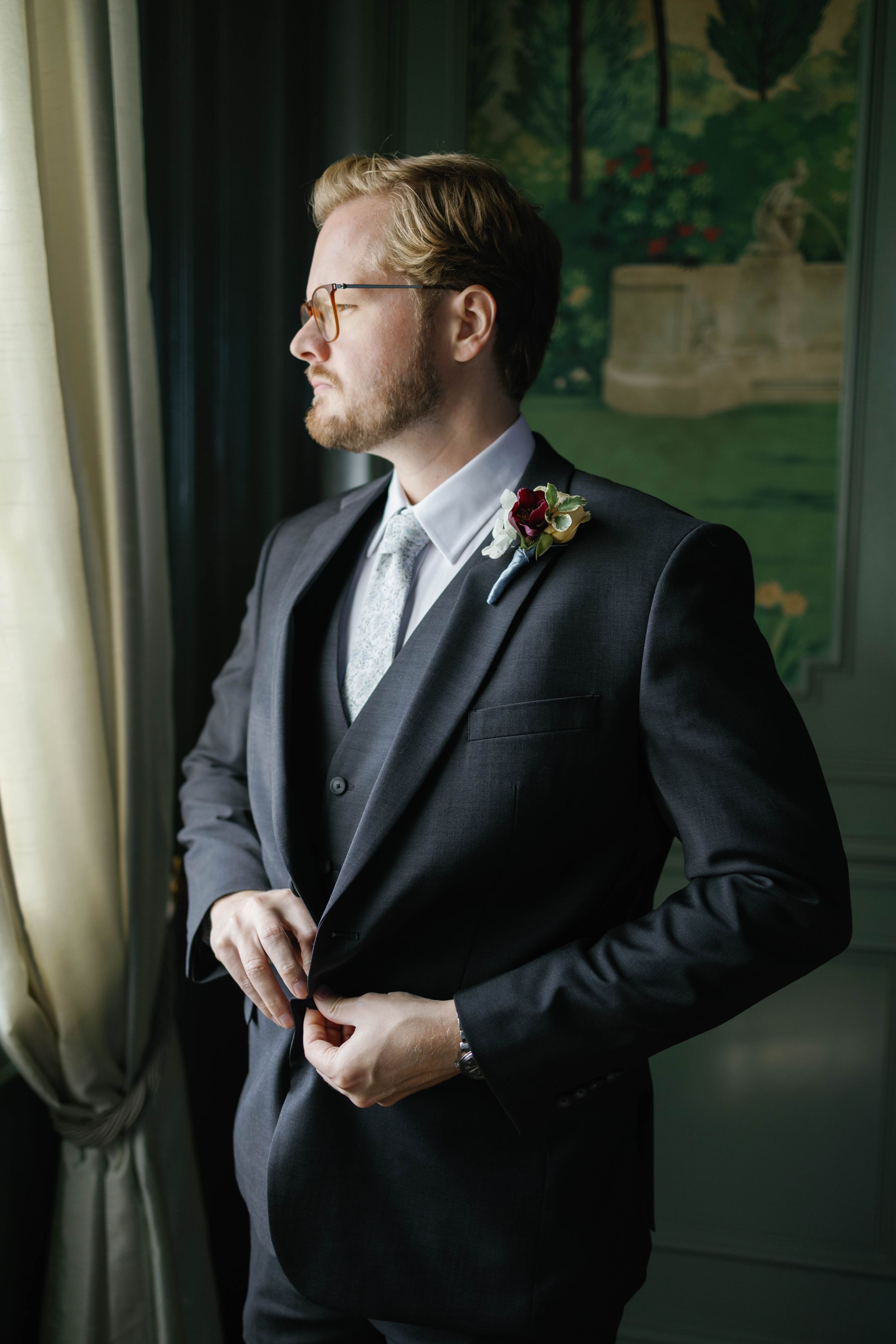 Dapper groom in dark suit with boutonniere buttoning jacket while gazing out window on wedding day.