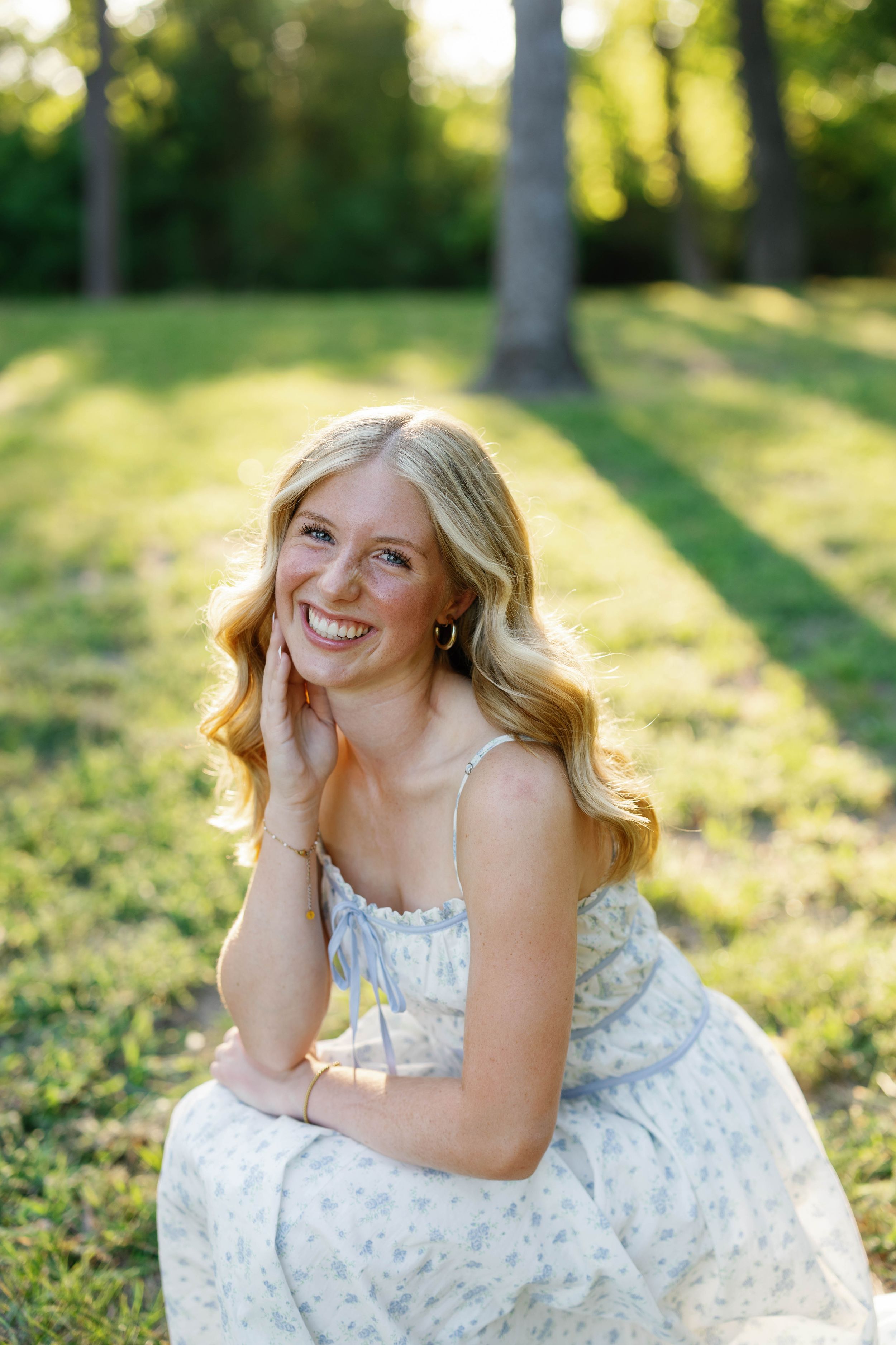 Smiling blonde woman in floral sundress sitting outdoors in sunlit green park setting.