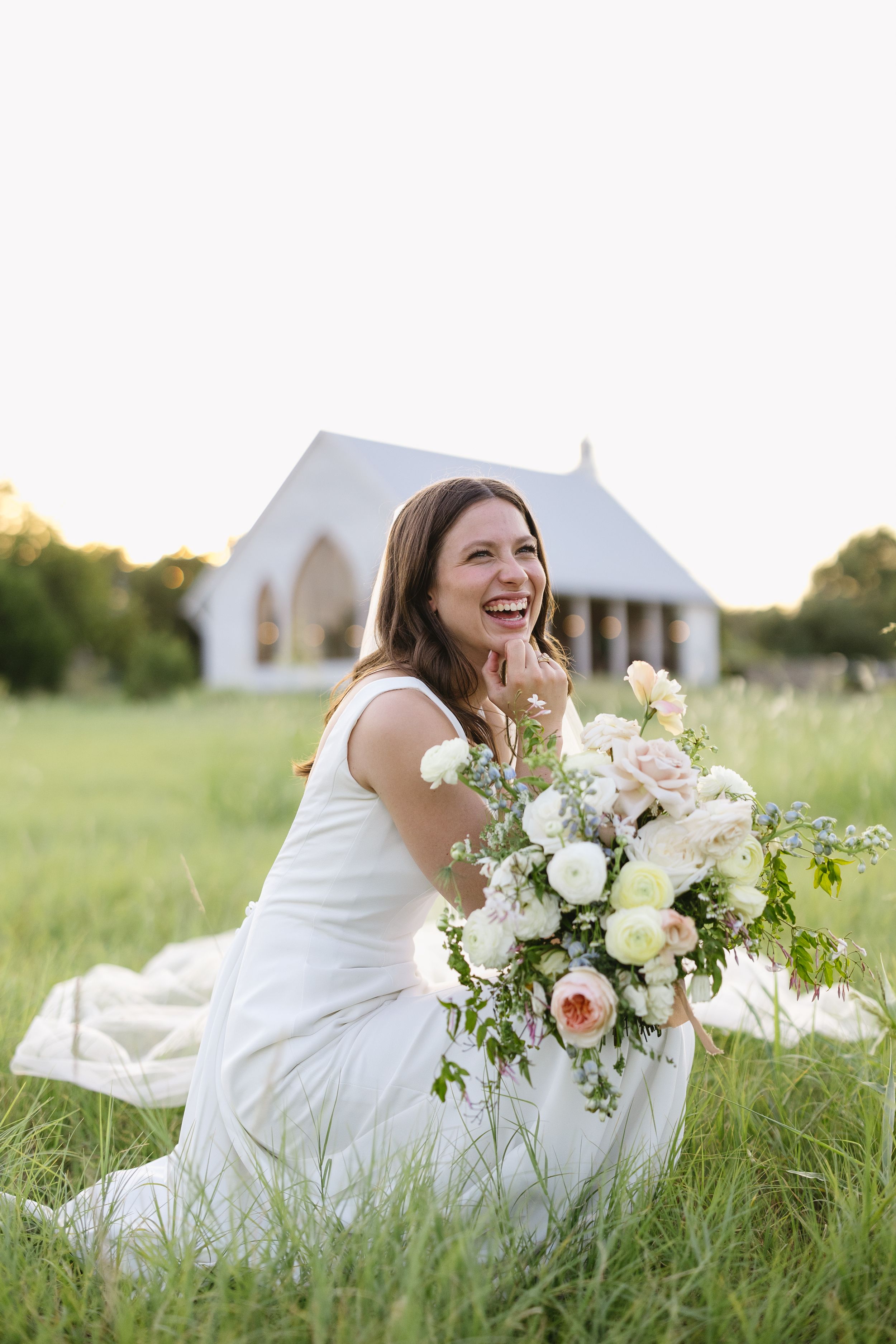 Joyful bride in white dress kneels in green meadow holding bouquet of white and blush roses near chapel.