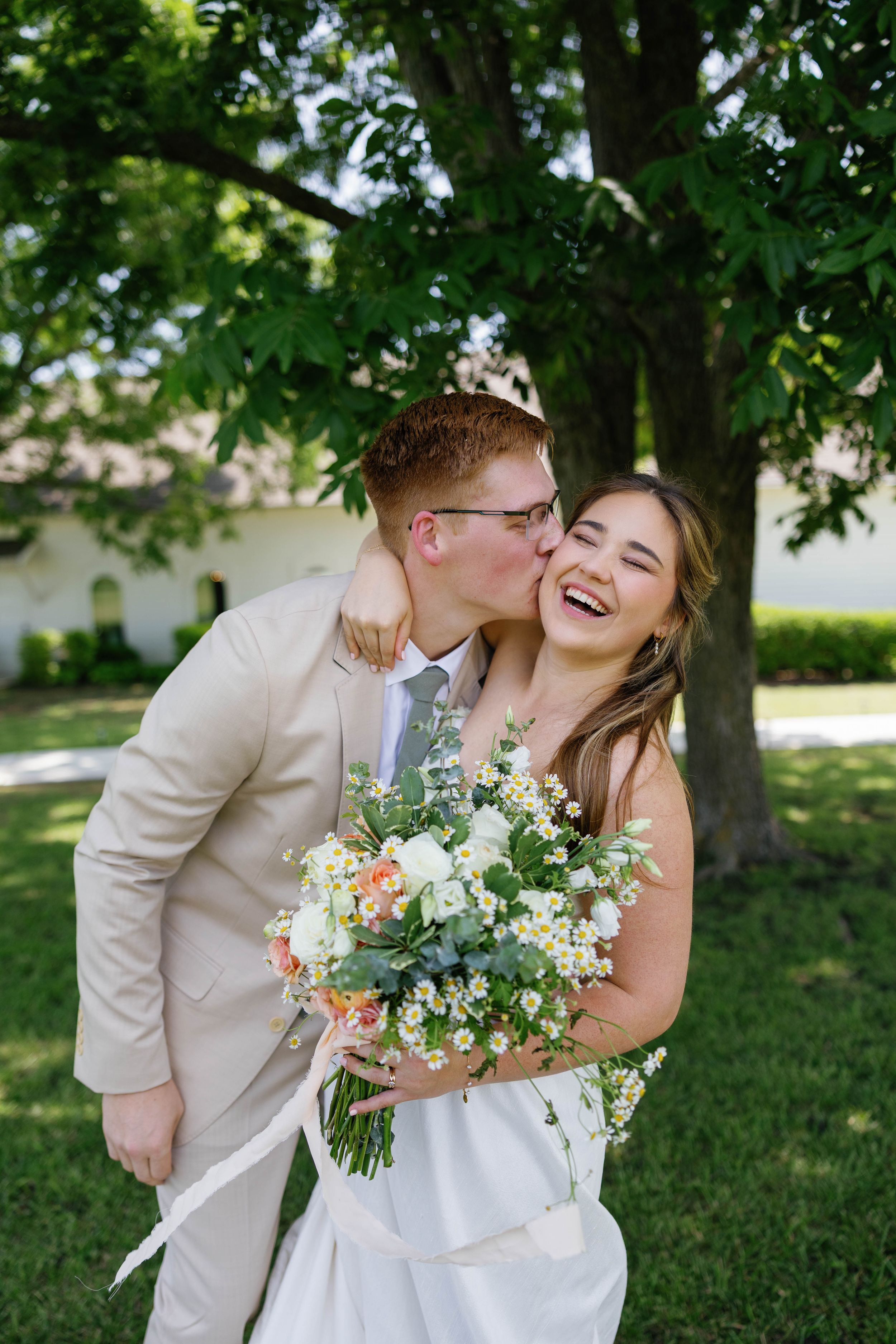 Joyful newlyweds sharing a kiss outdoors, bride laughing while holding white and peach wedding bouquet.