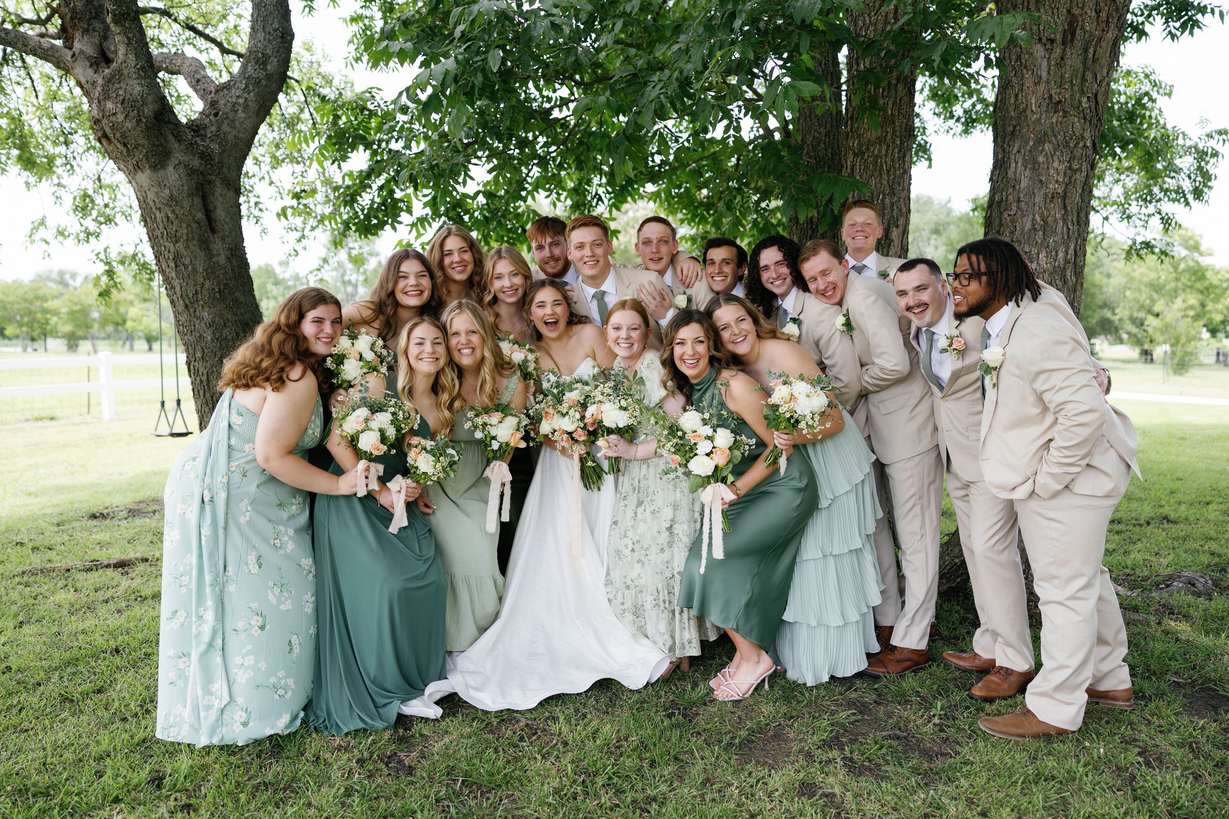Wedding party in sage green bridesmaid dresses and tan suits posing together under trees outdoors.