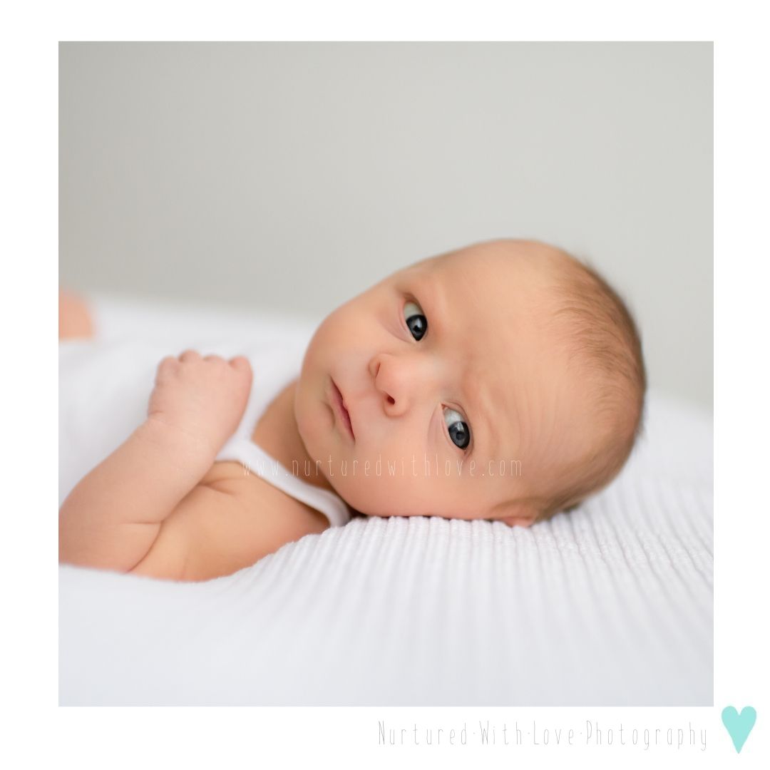 Sweet baby in white onesie lying on textured white blanket looking up with bright curious eyes.