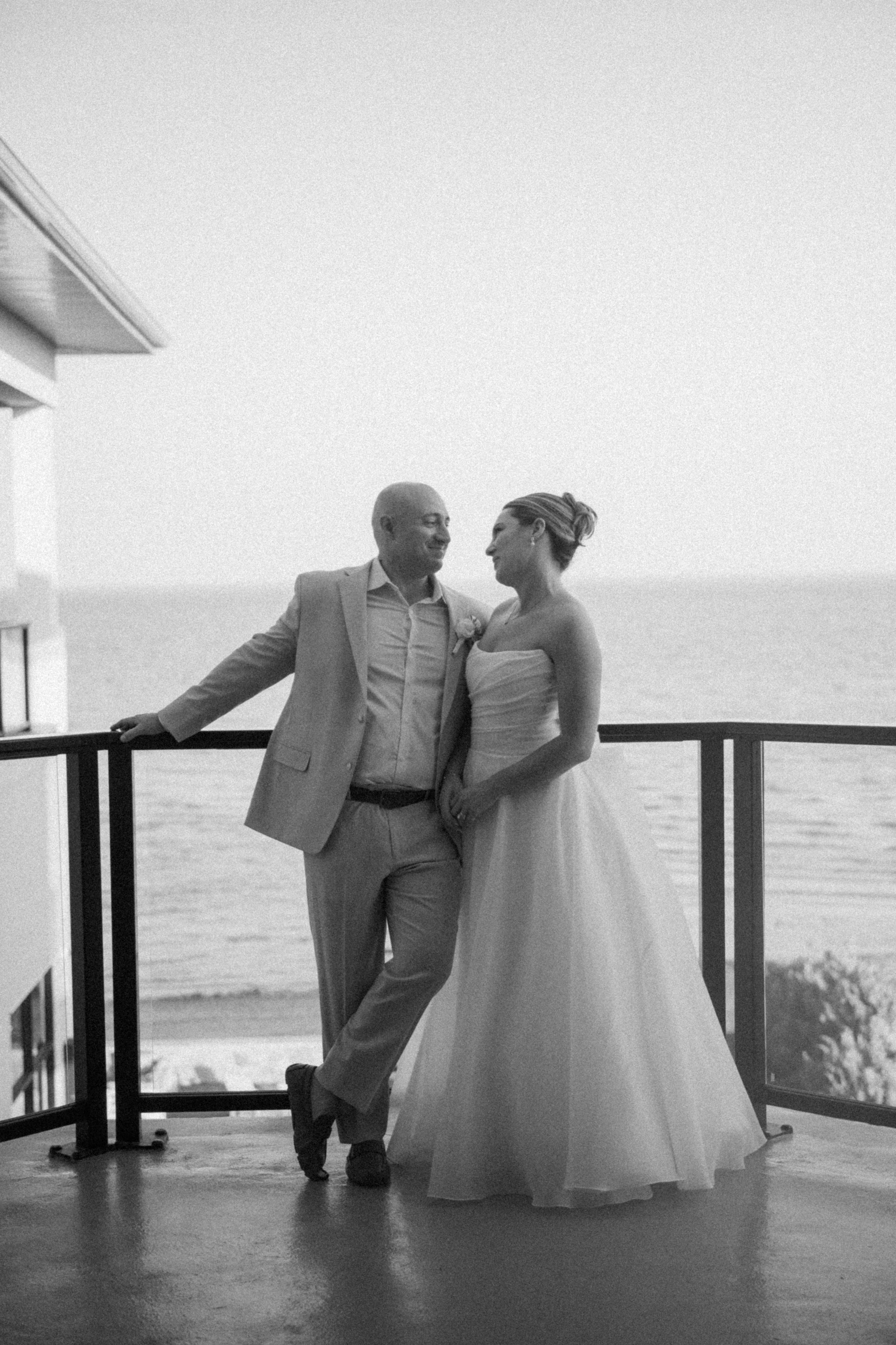 Black and white photo of a bride and groom sharing a romantic moment on a seaside balcony on their wedding day.