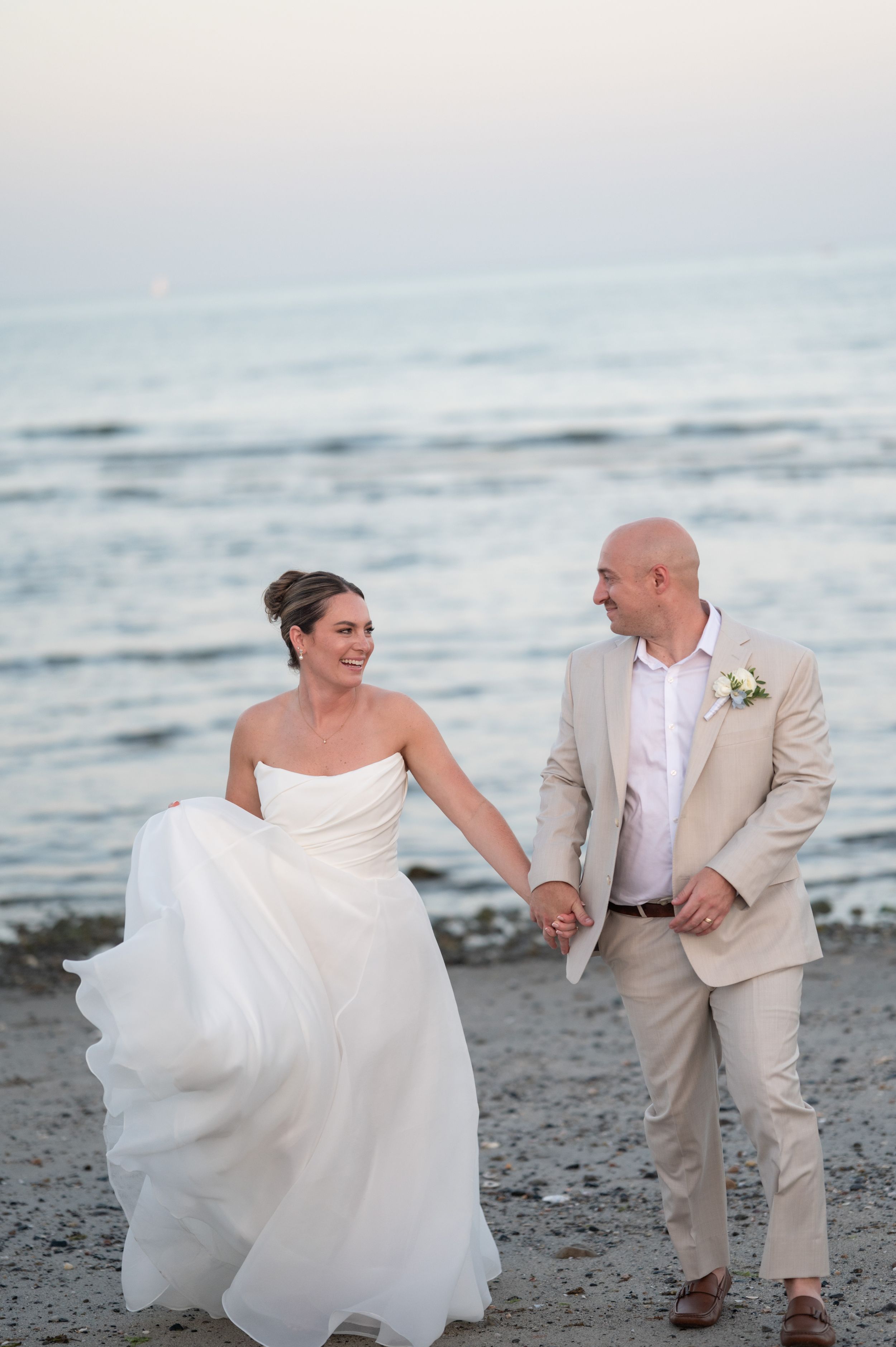 Bride in white strapless gown and groom in tan suit hold hands walking on beach at sunset.