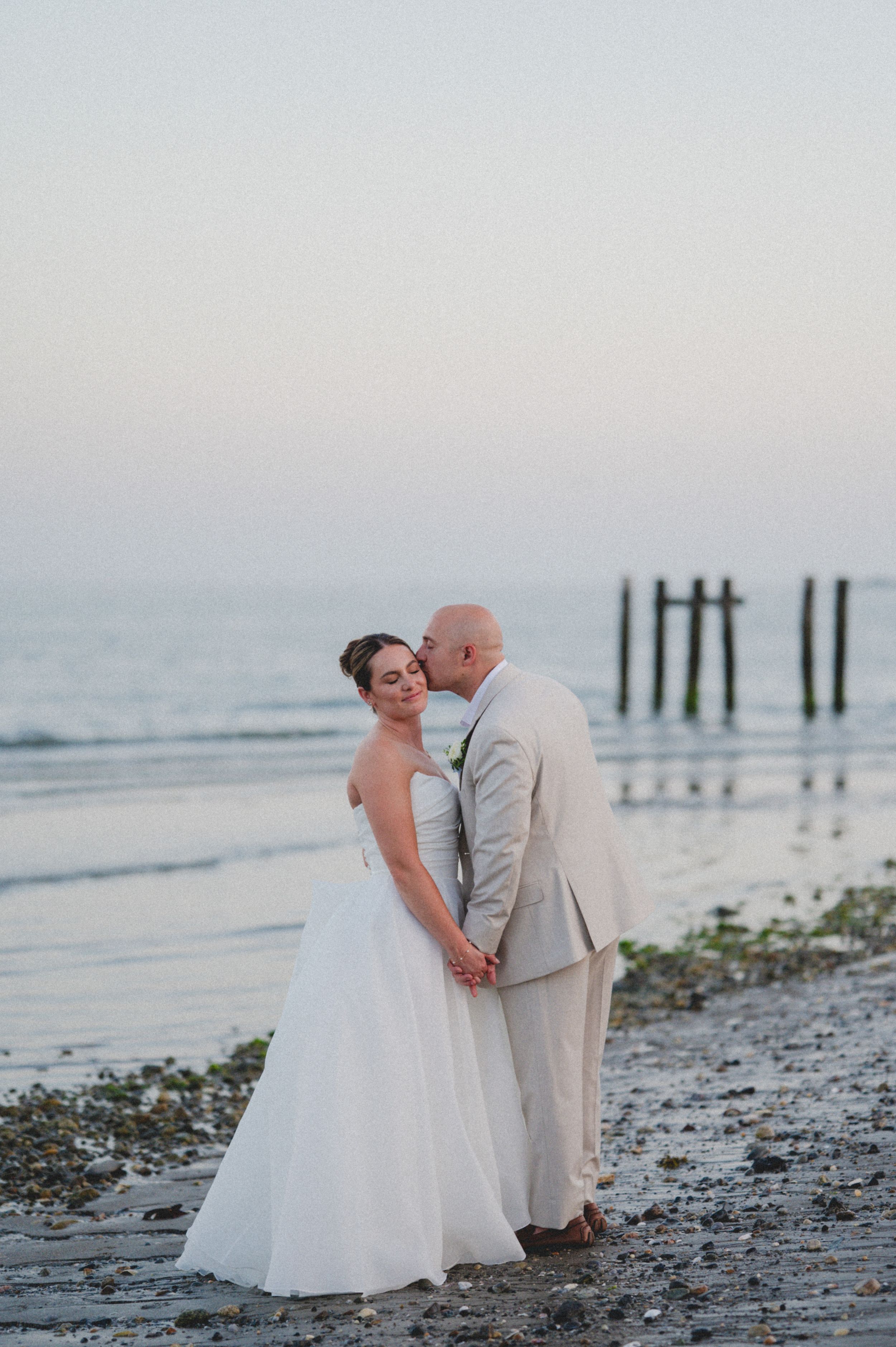 Bride and groom share a romantic kiss on a beach at sunset, with the groom in a tan suit and bride in a white gown.
