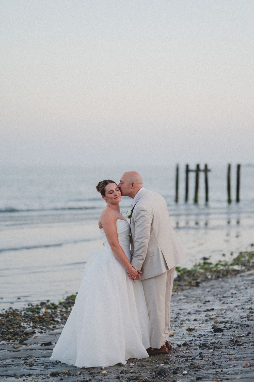 Bride and groom share a romantic kiss on a beach at sunset, with the groom in a tan suit and bride in a white gown.