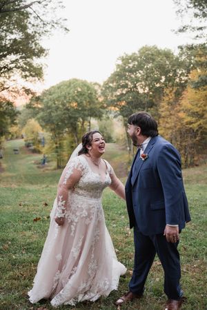 A laughing bride in lace gown and groom in navy suit share a joyful moment outdoors amid autumn foliage.