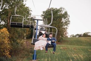 Bride and groom ride a ski lift together on a green hillside, celebrating their wedding day outdoors.