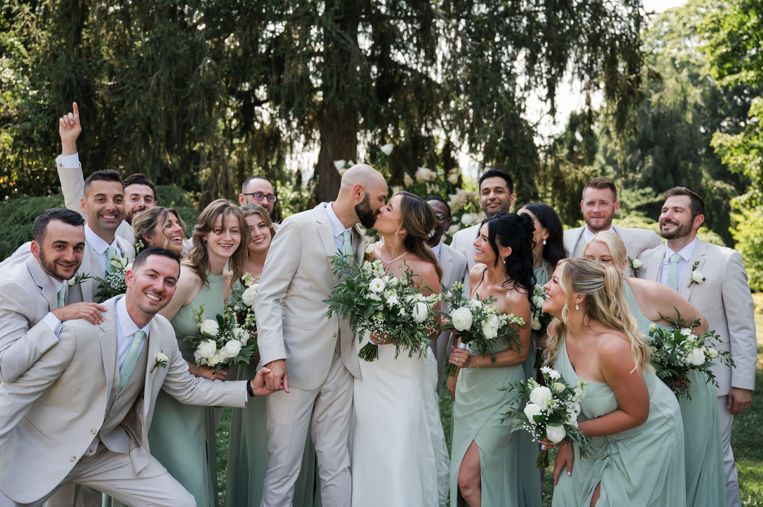 Wedding party in light colored attire poses joyfully together under trees at an outdoor celebration at Kalmia Garden at Gastler Farm