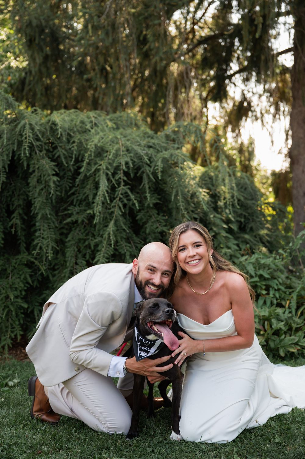 A couple kneels with their black dog against a backdrop of drooping evergreen trees. Kalmia Garden at Gastler Farm