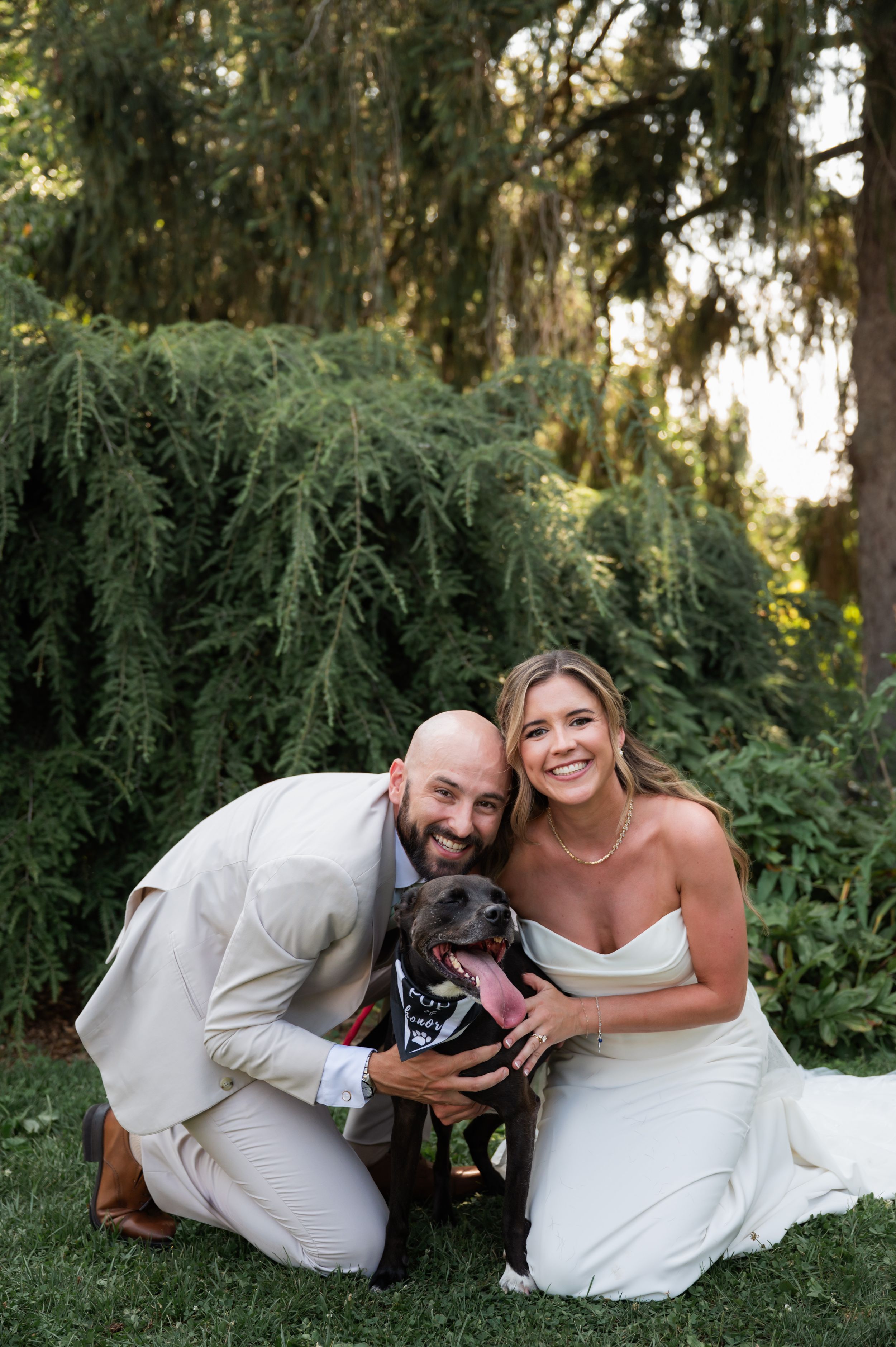 A couple kneels with their black dog against a backdrop of drooping evergreen trees. Kalmia Garden at Gastler Farm