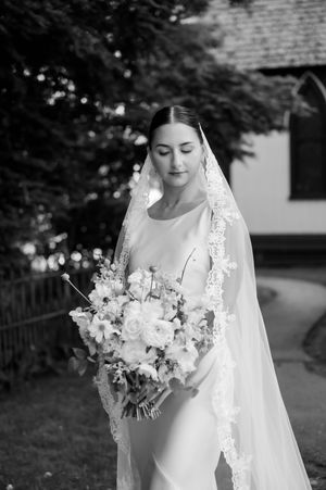Elegant bride in lace veil holds white floral bouquet in outdoor garden setting. pequot chapel