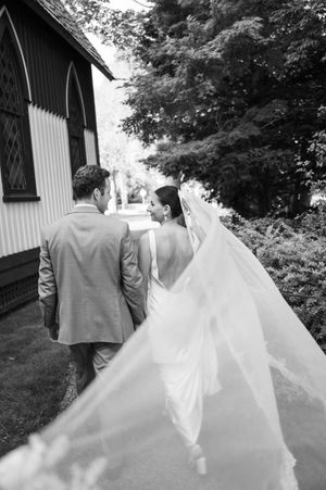 Flowing white wedding dress train follows couple walking through garden path in black and white. pequot chapel