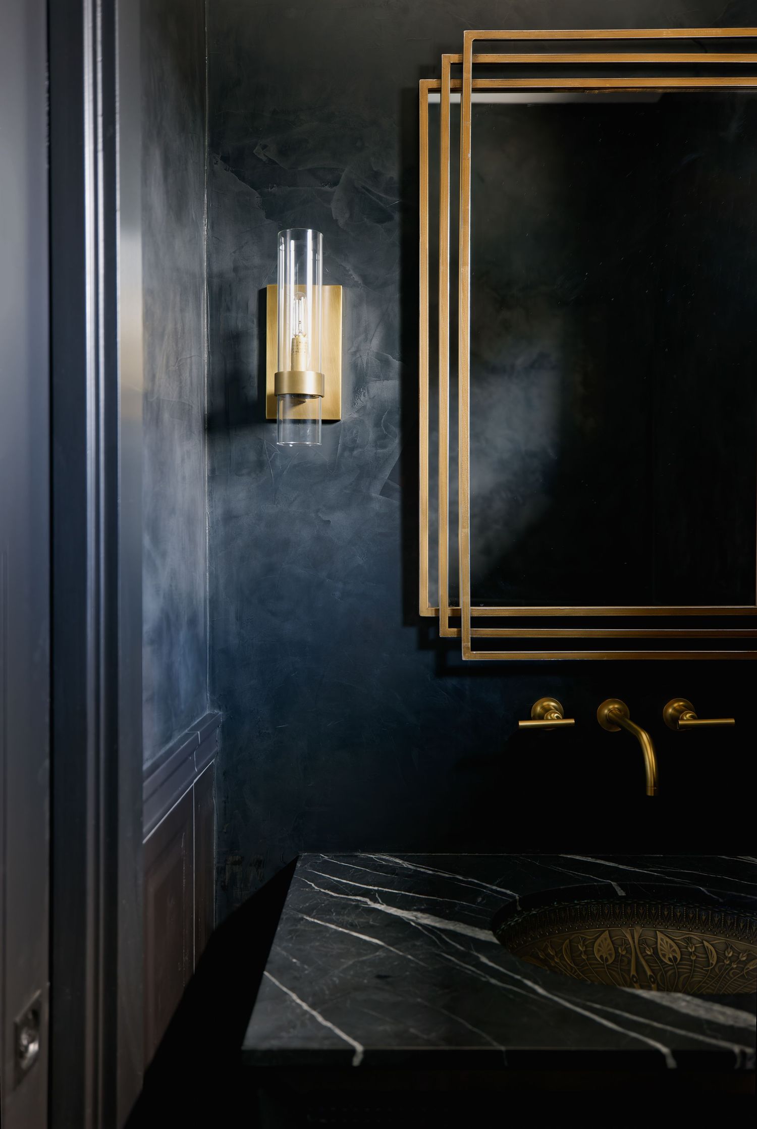 Dark moody bathroom with black marble sink, gold brass fixtures, ornate mirror frame, and elegant wall sconce lighting.