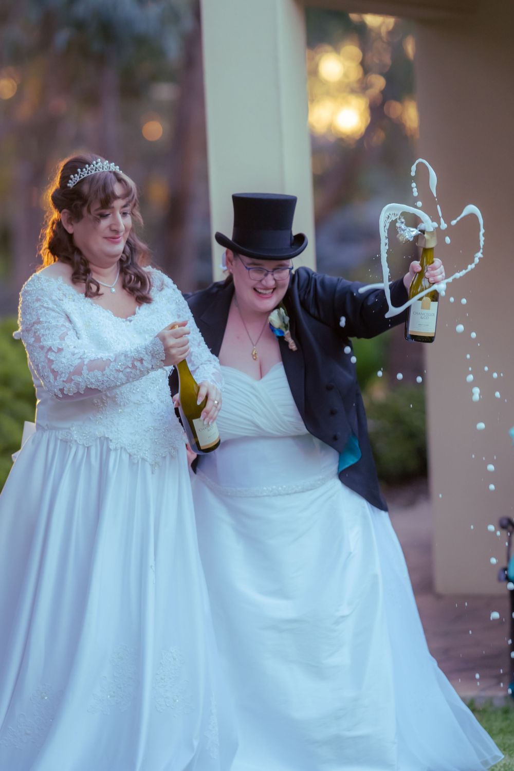 Two lesbian brides in wedding attire joyfully spray champagne outdoors with bokeh lights in the background.