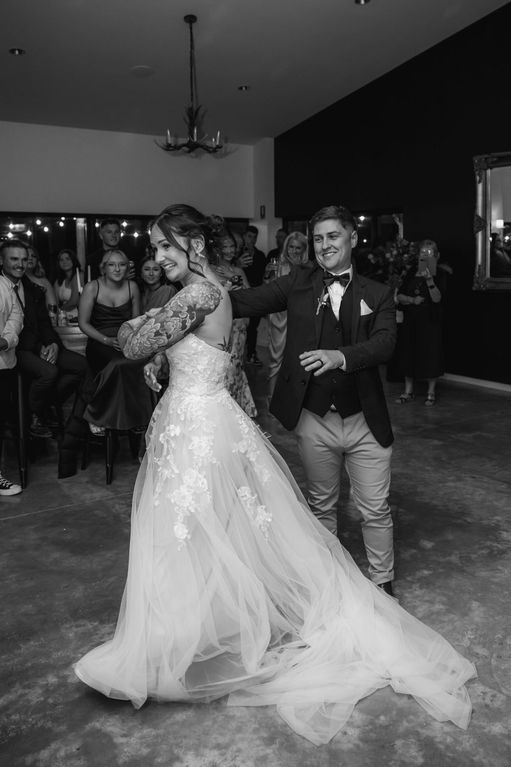 A couple performs their first dance at a wedding reception with guests looking on in black and white photography.