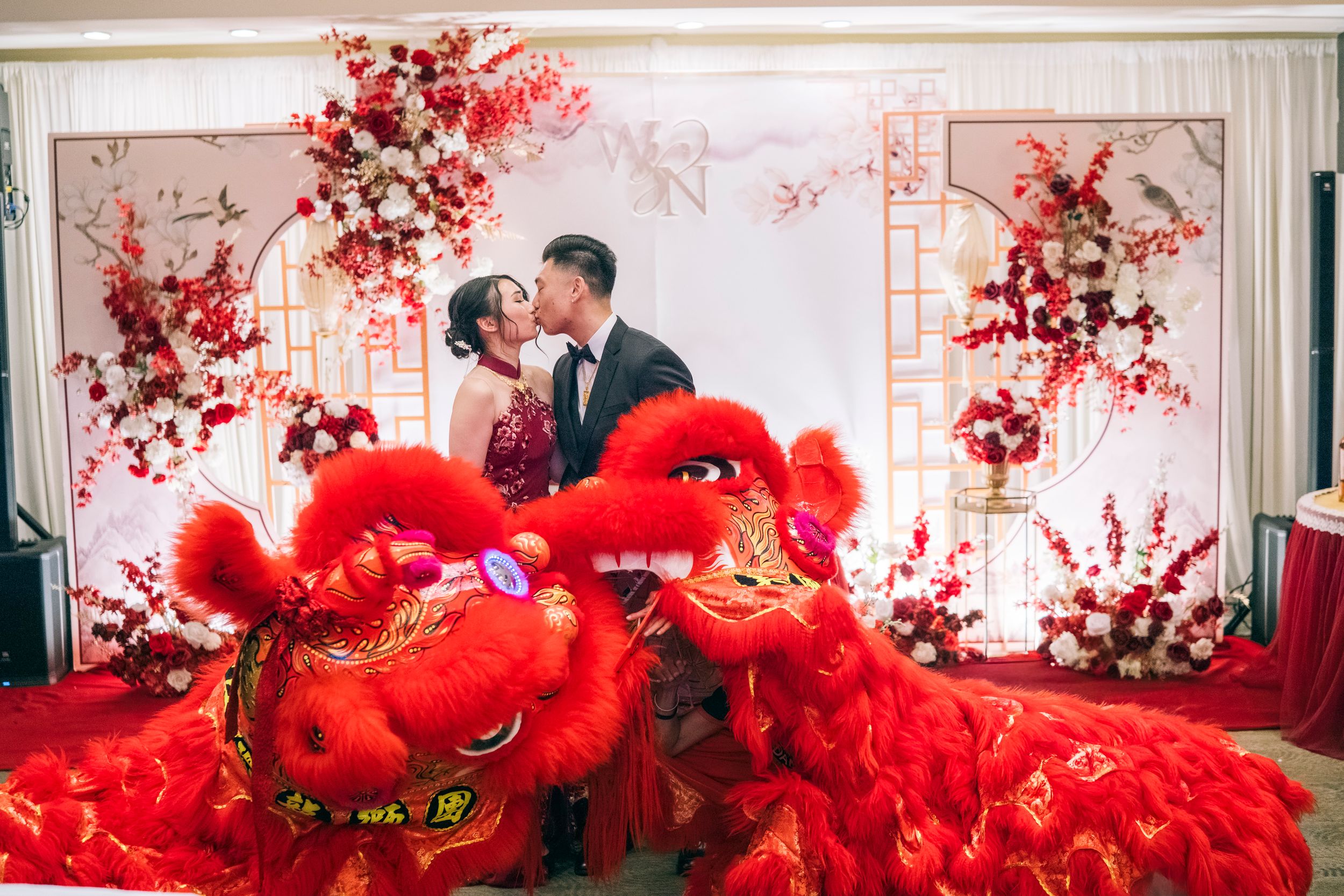 Couple kissing at Chinese wedding reception with red lion dance costume and floral arch backdrop.