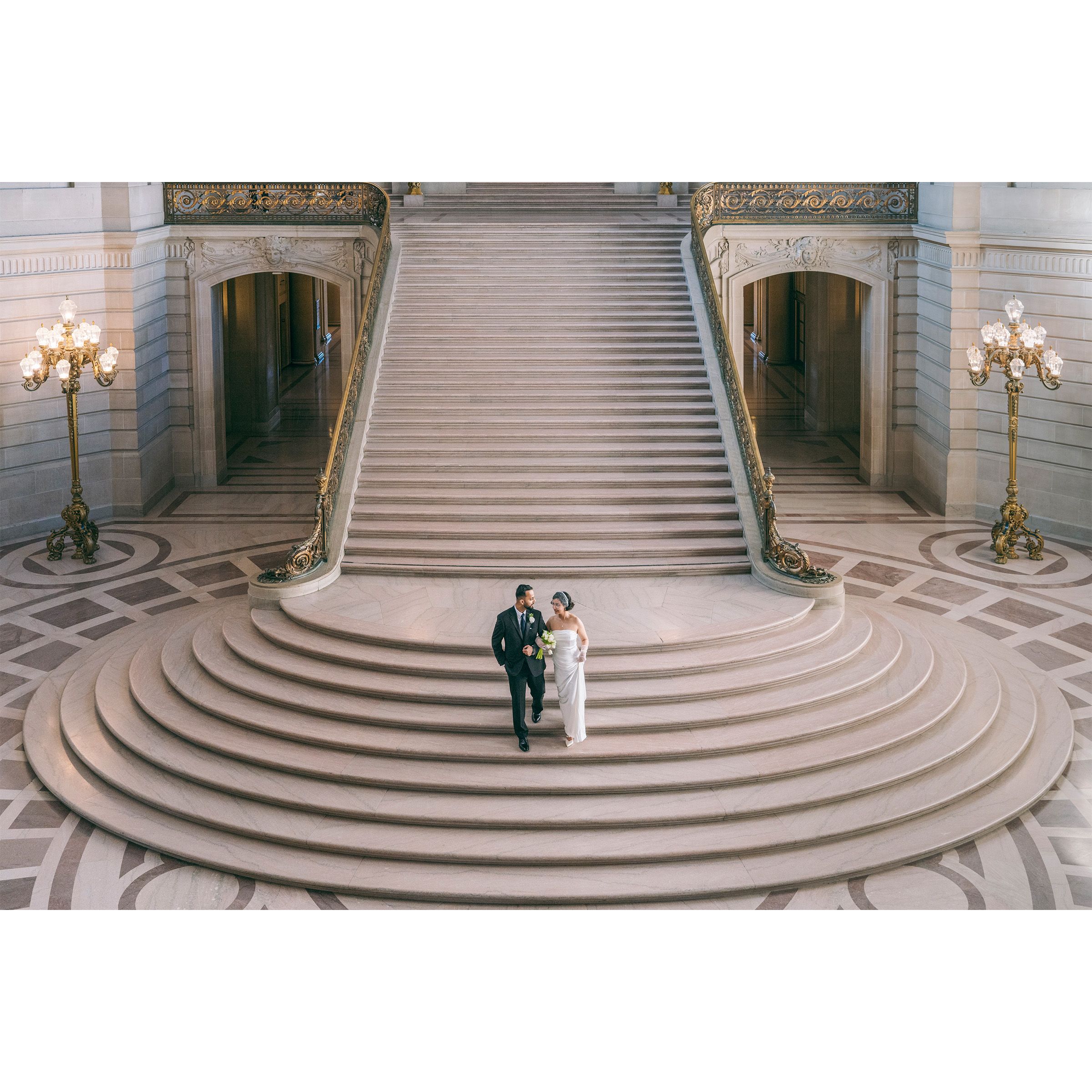 Wedding couple walking hand-in-hand on grand marble staircase at San Francisco City Hall from aerial view.