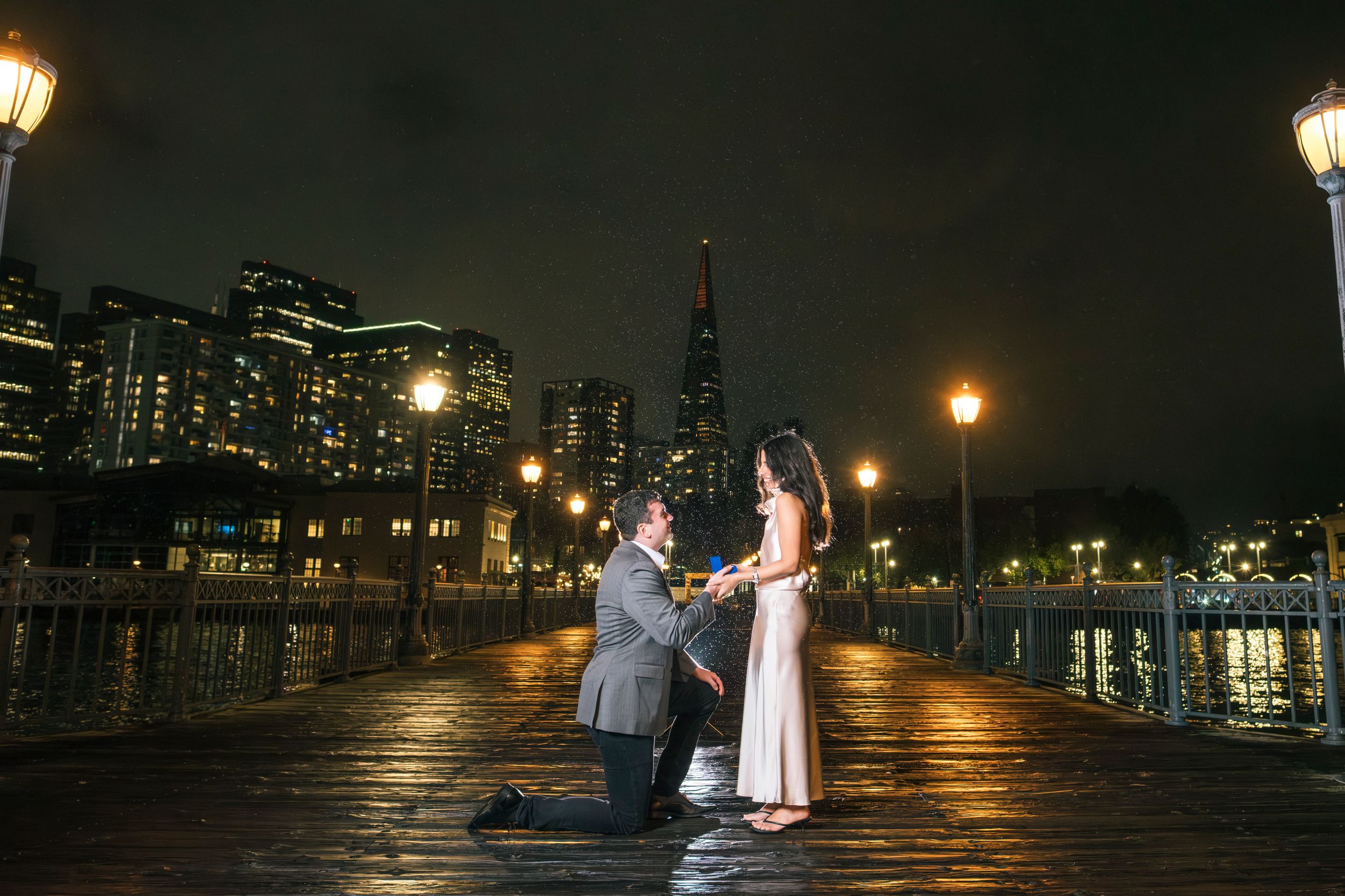 Man proposing on one knee on a waterfront pier at night with city skyline lights reflecting on wet dock.
