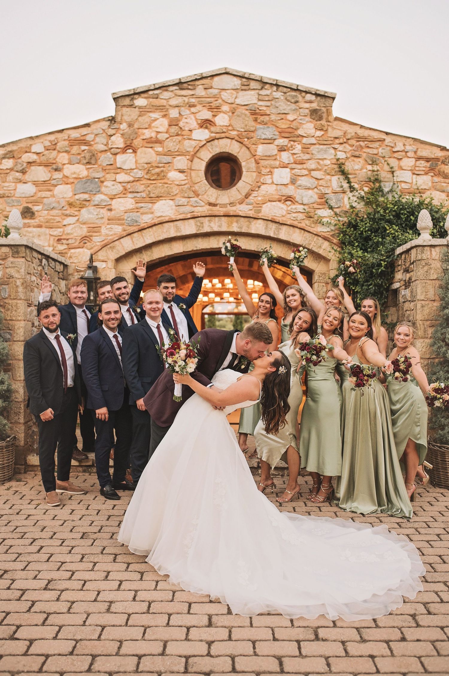 Wedding party celebrates outside stone church with bridal party in sage green dresses and groomsmen in dark suits.
