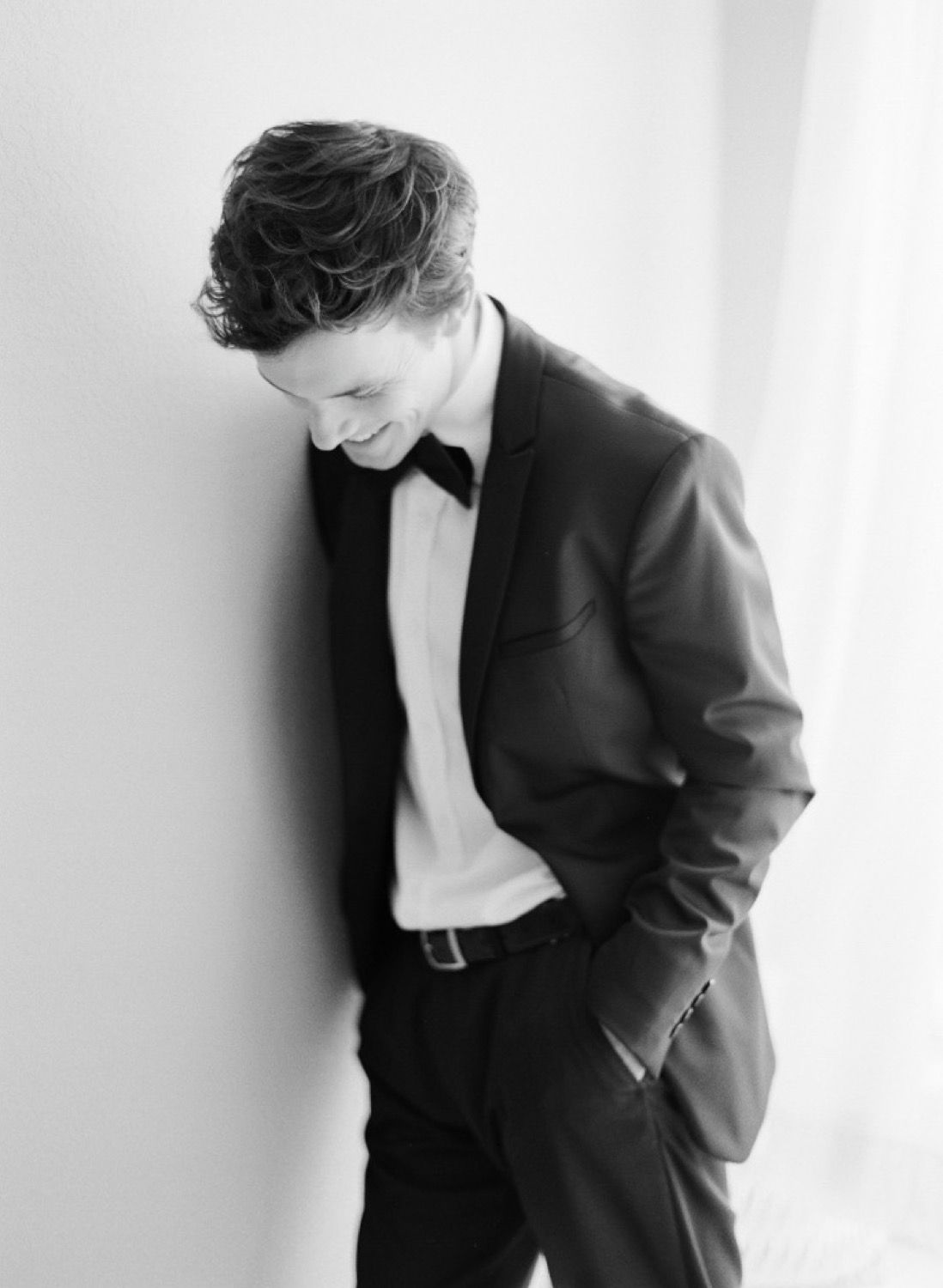Smiling groom in black tuxedo with bow tie leans against white wall in elegant black and white portrait.
