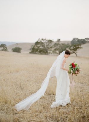 Bride in white strapless gown with cathedral veil holds colorful bouquet in golden California meadow.