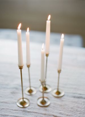 Five elegant white taper candles in slim gold candlestick holders glowing on a draped table.