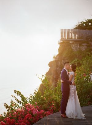 Couple in wedding attire sharing a romantic moment on a cliffside deck surrounded by tropical flowers at sunset.
