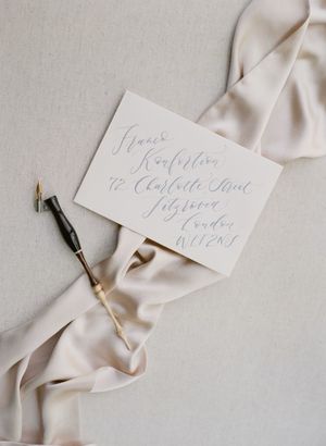 Minimalist wedding envelope with soft gray calligraphy address resting on ivory silk ribbon with a dip pen.