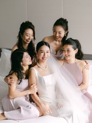 Bride in white gown with veil surrounded by four bridesmaids in pink dresses on a bed.