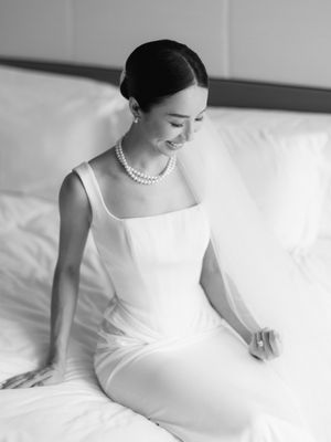 Elegant bride in white sleeveless gown and pearl necklace sitting on bed, smiling in black and white photo.
