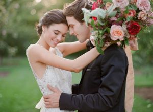 Romantic bride and groom embracing near vibrant red and pink floral wedding bouquet.