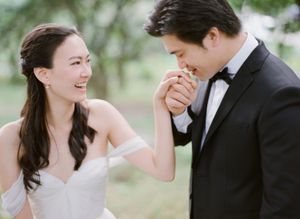 Asian bride and groom sharing romantic moment as groom kisses bride's hand outdoors.